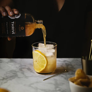 A hand is pouring a bottle of THRIVE, Manuka Mana's turkey tail tonic, into a glass with ice.