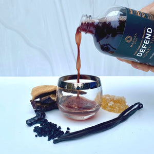 A hand is pouring Reishi Defend tonic into a cup surrounded by ingredients.
