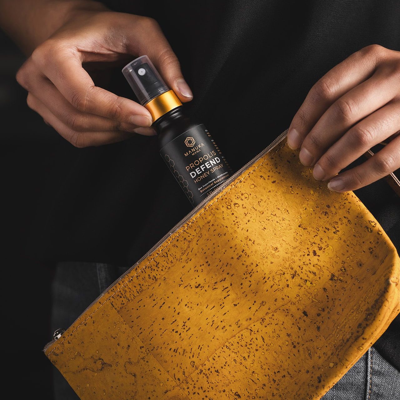 A person's hands holding a small black spray bottle labeled 'Manuka Mana Propolis Defend Spray' next to a textured mustard yellow pouch against a dark background.