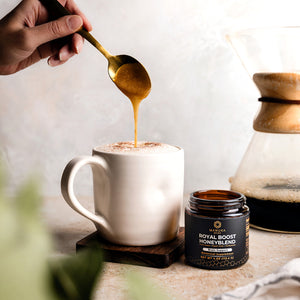 A jar of the Manuka Mana Royal Boost Honeyblend is pictured next to a cup of coffee that has a spoon dripping the honeyblend into it. 