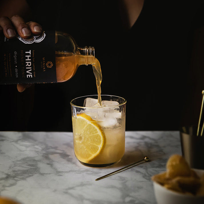 A person pours an amber liquid from a bottle labeled THRIVE into a clear glass containing ice and a lemon slice, set on a marble tabletop with blurred lemons and a small bowl in the foreground.