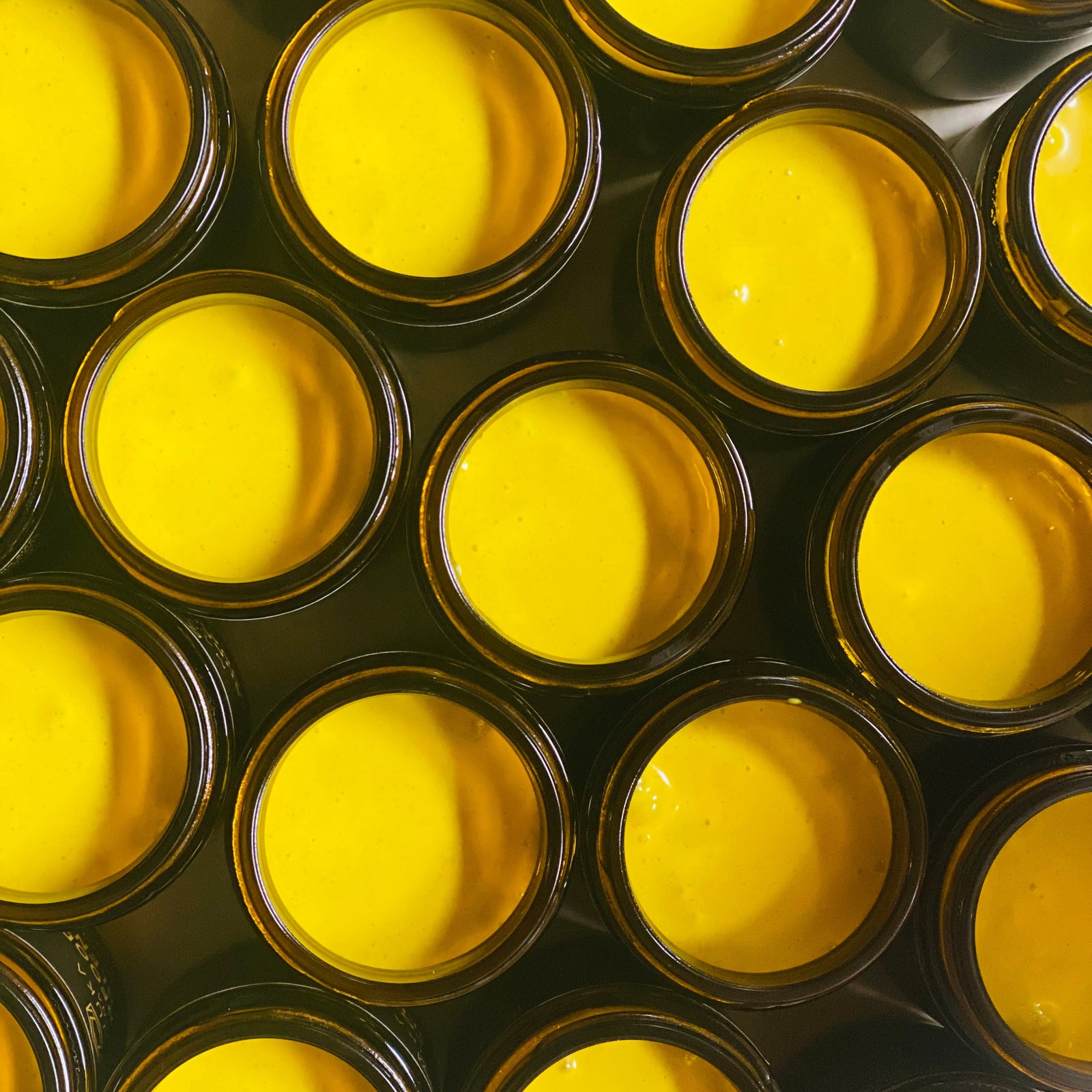 Multiple amber glass jars filled with bright yellow cream or balm, arranged closely together in a repeating pattern.